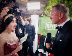 An actress interviews a male celebrity on the red carpet with cameras and crew in the background.