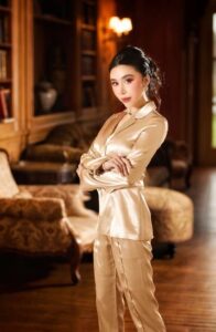 A woman in elegant gold satin pajamas stands in a luxurious room.